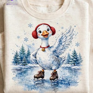 Puede incluir: Una sudadera de color crema con una ilustraci&oacute;n en acuarela de un ganso blanco que lleva orejeras rojas y un collar rojo con un cascabel dorado. El ganso patina sobre un estanque helado rodeado de pinos y copos de nieve que caen.