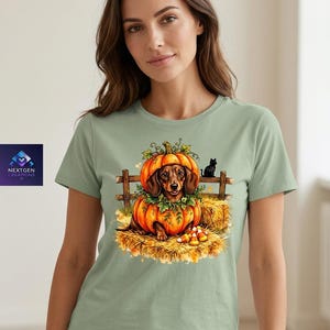 May include: A sage green t-shirt featuring a graphic print of a brown dachshund sitting inside a large orange pumpkin. The scene includes a wooden fence, a small black cat, hay bales, and candy corn. The logo NEXTGEN CREATIONS is visible.