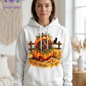 May include: A white hooded sweatshirt featuring a graphic print of a brown dachshund sitting inside a large orange pumpkin. The scene includes a wooden fence, hay bales, green leaves, candy corn, and a small black cat silhouette.
