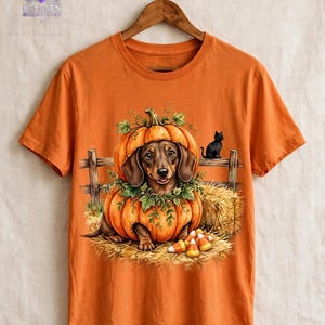 May include: An orange short-sleeved t-shirt featuring a graphic of a brown dachshund sitting inside a large pumpkin. The scene includes a wooden fence, hay, a small black cat, and candy corn. The shirt hangs on a wooden hanger.