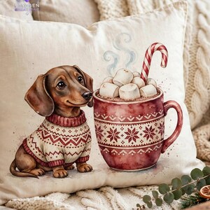 May include: A cream-colored throw pillow featuring a watercolor illustration of a brown dachshund wearing a cream and red patterned sweater sitting next to a large red mug filled with hot cocoa, white marshmallows, and a candy cane.