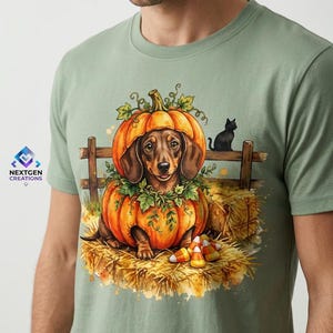 May include: A sage green t-shirt featuring a graphic of a brown dachshund sitting inside a carved orange pumpkin. The scene includes a wooden fence, hay bales, a black cat, and candy corn. The text NEXTGEN CREATIONS is printed on the left.