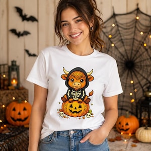 May include: A white short-sleeved t-shirt featuring a graphic print of a cartoon Highland cow wearing a black skeleton hoodie and sitting on top of a carved jack-o'-lantern pumpkin, surrounded by small skulls and candy corn illustrations.