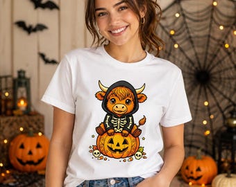 Puede incluir: Una camiseta blanca de manga corta con un estampado gráfico de un ternero Highland de dibujos animados con una sudadera negra de esqueleto, sentado sobre una calabaza de Halloween tallada, rodeado de pequeñas calaveras y dulces.