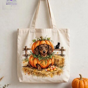 May include: A beige canvas tote bag featuring a printed illustration of a brown dachshund sitting inside a large orange pumpkin. The scene includes a small black cat on a wooden fence, hay bales, and candy corn on the ground.