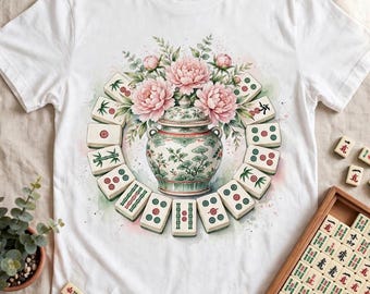 Puede incluir: Una camiseta blanca con un gráfico central de una vasija de cerámica decorativa rodeada por una disposición circular de fichas de Mahjong y flores de peonía rosas, con detalles de estilo acuarela en tonos verdes y rosas.