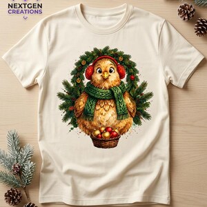 K&ouml;nnte beinhalten: Ein cremefarbenes T-Shirt mit einer Grafik eines flauschigen braunen Vogels, der rote Ohrensch&uuml;tzer und einen gr&uuml;nen Strickschal tr&auml;gt. Der Vogel ist in einem Tannenkranz zentriert und h&auml;lt einen braunen Korb mit roten und goldenen Weihnachtskugeln.