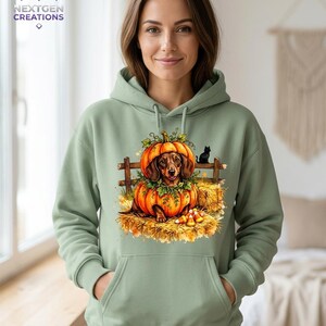 May include: A sage green hooded sweatshirt featuring a graphic print of a brown dachshund sitting inside a large orange pumpkin. The design includes a wooden fence, hay bales, candy corn, and a small black cat silhouette.