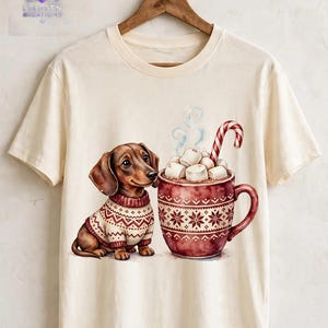 May include: A cream-colored t-shirt featuring a graphic of a brown dachshund wearing a patterned sweater sitting next to a large red mug filled with hot cocoa, marshmallows, and a candy cane. The mug has a white snowflake-like pattern.