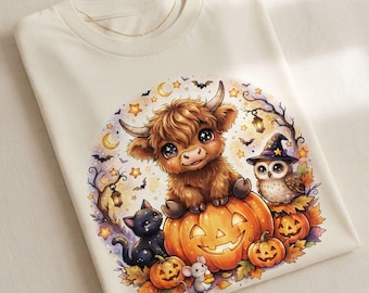 Puede incluir: Una camiseta de color crema con un gráfico de una vaca highland peluda sentada sobre una calabaza grande, acompañada por un gato negro, un ratón pequeño con un dulce, y un búho con sombrero de bruja, rodeados de estrellas y murciélagos.