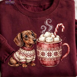 May include: A maroon crewneck sweatshirt featuring a graphic of a brown dachshund wearing a white and maroon patterned sweater next to a mug of hot cocoa topped with marshmallows and a candy cane. The text NEXTGEN CREATIONS is visible.