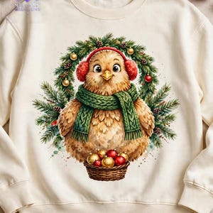K&ouml;nnte beinhalten: Ein cremefarbenes Sweatshirt mit Rundhalsausschnitt und einem Grafikdruck eines flauschigen braunen Vogels mit roten Ohrensch&uuml;tzern und einem gr&uuml;nen Strickschal. Der Vogel sitzt in einem Tannenkranz und h&auml;lt einen Korb mit roten und goldenen Kugeln.