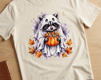 Puede incluir: Una camiseta de manga corta color crema con un gráfico de un mapache vestido con un disfraz de fantasma blanco. El mapache sostiene una calabaza naranja llena de dulces, rodeado de hojas de otoño naranjas y amarillas dispersas.