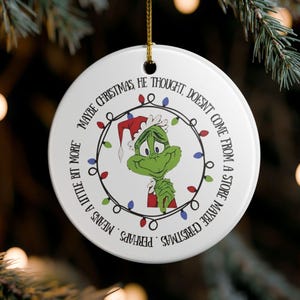 Könnte beinhalten: Weißer, runder Weihnachtsschmuck mit dem Grinch mit Weihnachtsmütze, umgeben von einer Lichterkette. Der Text lautet: „Vielleicht kommt Weihnachten, dachte er, nicht aus einem Geschäft. Vielleicht bedeutet Weihnachten ein bisschen mehr.“