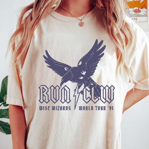 May include: Cream-colored t-shirt with a dark blue raven graphic and the text "RUN CLAW" above "WISE WIZARDS WORLD TOUR '91". The shirt has a relaxed fit and short sleeves. The design features a lightning bolt.