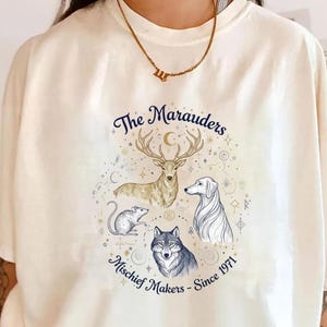 May include: Cream-colored t-shirt featuring a circular design with a deer, dog, rat, and wolf illustration. The text reads "The Marauders" and "Mischief Makers - Since 1971" in navy blue script. The shirt has short sleeves.