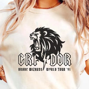 May include: Cream-colored t-shirt featuring a black graphic of a roaring lion's head with the word "GRIFFINDOR" and "BRAVE WIZARDS WORLD TOUR '91" printed below. The design includes a lightning bolt.