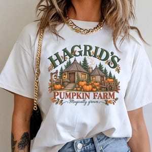 Può includere: Una t-shirt bianca a maniche corte con una grafica raffigurante un cottage in pietra circondato da pini e zucche. Il testo recita HAGRID'S PUMPKIN FARM Magically grown in lettere verdi e arancioni con foglie autunnali e un carretto.