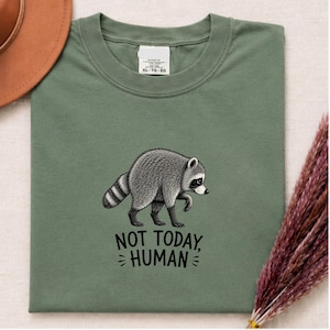 May include: An olive green crew neck t-shirt featuring a black and white illustration of a raccoon walking. Below the raccoon, the text reads NOT TODAY, HUMAN in a bold, black, sans-serif font. The shirt has a white interior neck label.