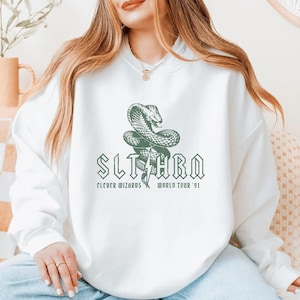 May include: White sweatshirt featuring a green snake graphic with the word "SLITHRA" and the text "Clever Wizards World Tour '91". The design is centered on the front of the sweatshirt.