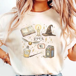 Puede incluir: Camiseta color crema con ilustraciones de un libro, una vela, un sombrero seleccionador, gafas, una snitch dorada, una botella de poción, una varita, un billete 9 3/4 y un cofre del tesoro. El diseño incluye estrellas y rayos.