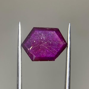 May include: A faceted hexagonal gemstone with a deep magenta and purple hue, featuring visible internal inclusions and surface fractures, held securely between the two pointed metal tips of a pair of silver-toned professional jeweler tweezers.