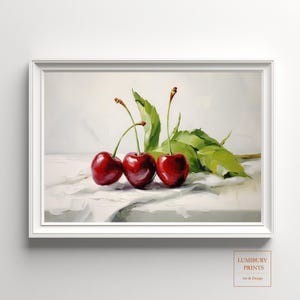 May include: A framed still life painting featuring three ripe, red cherries with green leaves and stems. The cherries are arranged on a white surface, with a soft, neutral background. The artwork is signed "LUMIBURY PRINTS Art & Design".