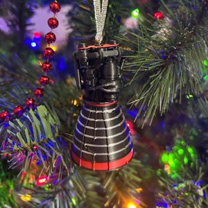 May include: A black and red rocket engine Christmas ornament with silver accents. The ornament is hanging on a green Christmas tree with red bead garland and colorful lights.
