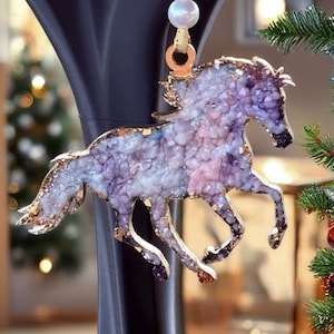 May include: A horse-shaped ornament featuring a textured, crystalline surface in shades of purple, pink, and white. The edges are outlined in a metallic gold finish. It hangs from a gold ribbon accented with a single white pearl-like bead.