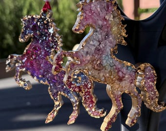 May include: Two horse-shaped ornaments featuring textured, crystalline surfaces with gold-painted edges. One horse displays deep purple and magenta tones, while the other features a gradient of pink, yellow, and blue. Each hangs from a pink ribbon with a white bead.