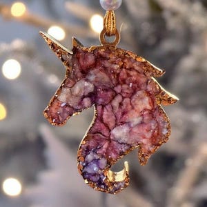 May include: A unicorn-shaped decoration with a gold-coloured outline and a textured, multi-coloured interior. The decoration features shades of purple, pink, and white, and is suspended by a gold ribbon and a small, pearlescent bead. The background is blurred with soft, out-of-focus lights.