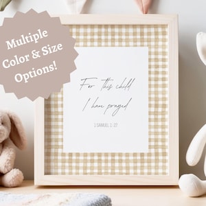 Può includere: Stampa incorniciata con il testo "For this child I have prayed" e il versetto biblico 1 Samuele 1:27. La stampa ha un motivo a quadretti ed è circondata da una cornice in legno chiaro. Un coniglietto di peluche e un coniglietto di peluche sono su entrambi i lati.