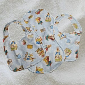 May include: Three baby bibs featuring a light blue pattern with illustrations of Peter Rabbit and books. The bibs are made of soft fabric and have a rounded shape with a neck opening. The bibs are designed for babies.
