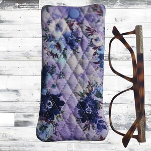 May include: A quilted glasses case with a floral design in purple and blue, fastened with a blue button. Beside it are brown tortoiseshell spectacles.