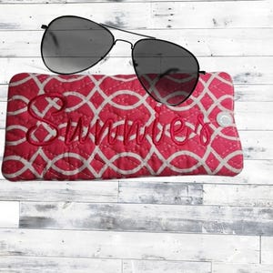 May include: Aviator sunglasses with black frames and dark lenses resting on a rectangular quilted fabric case. The case features a pink and white geometric pattern with the word Sunnies embroidered in pink script across the front surface.