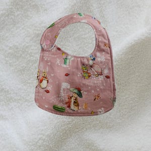 May include: A pink baby bib with a rounded top and a snap closure. The bib features a repeating pattern of illustrations of rabbits, ducks, and other garden elements. The bib is made of soft fabric and is designed to catch spills.