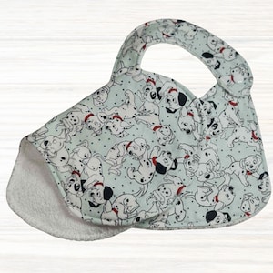 May include: A baby bib with a Dalmatian puppy print on a pale blue background. The bib has a curved shape with a handle and a soft, white terry cloth lining. The bib is designed for easy use.