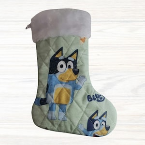 May include: A quilted Christmas stocking in light green featuring a repeating print of the cartoon character Bluey. The stocking has a white faux fur cuff at the top and includes the blue text logo Bluey printed on the fabric surface.