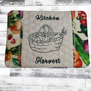 May include: Rectangular kitchen mat with a quilted center featuring a black embroidered basket filled with vegetables. The words "Kitchen Harvest" are above and below the basket. The edges have a colorful vegetable print.