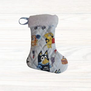 May include: A quilted light blue Christmas stocking featuring a repeating pattern of cartoon dogs, including one wearing an apron with the text World's Best Chef. The stocking has a white faux fur cuff and a small blue fabric hanging loop.