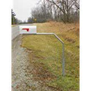 May include: A white mailbox with a red flag, mounted on a metal post beside a gravel road. The mailbox is rectangular and has a standard design. The setting is outdoors, with grass and trees in the background.