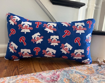 May include: A rectangular throw pillow featuring a repeating pattern of the Philadelphia Phillies baseball team logo and the word Phillies in white and red on a royal blue background. The design includes the iconic Liberty Bell graphic.