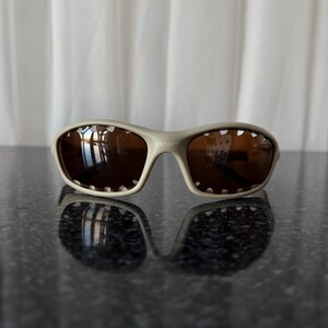 May include: Beige-framed sunglasses with brown lenses. The sunglasses have a sporty design with a curved frame and the word "SATISFY" printed on the frame. The lenses reflect the surrounding environment.