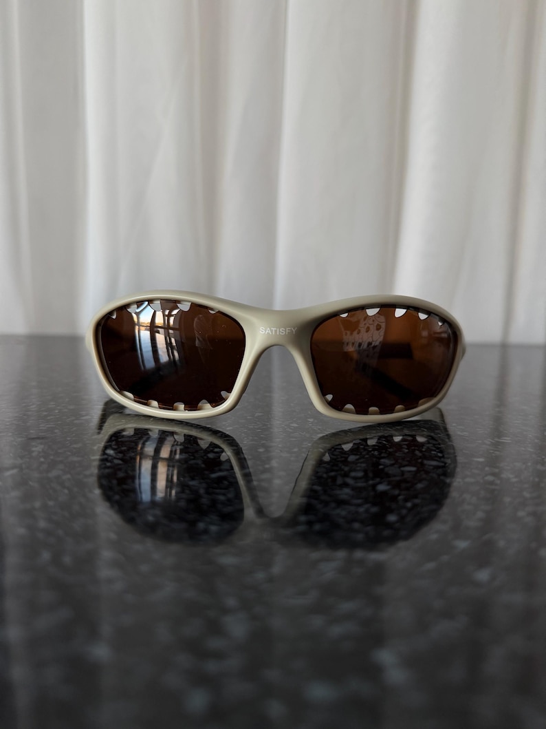 May include: Beige-framed sunglasses with brown lenses. The sunglasses have a sporty design with a curved frame and the word "SATISFY" printed on the frame. The lenses reflect the surrounding environment.