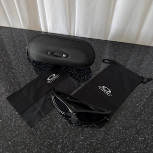 K&ouml;nnte beinhalten: Schwarze Sonnenbrille mit passendem Etui, Mikrofasertuch und Kordelzugbeutel. Die Sonnenbrille hat ein schlankes, modernes Design. Das ovale Etui ist strukturiert. Der Beutel und das Tuch tragen das Markenlogo.