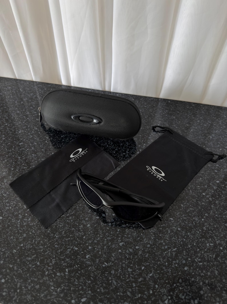 K&ouml;nnte beinhalten: Schwarze Sonnenbrille mit passendem Etui, Mikrofasertuch und Kordelzugbeutel. Die Sonnenbrille hat ein schlankes, modernes Design. Das ovale Etui ist strukturiert. Der Beutel und das Tuch tragen das Markenlogo.