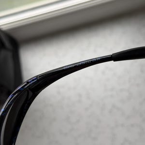 K&ouml;nnte beinhalten: Schwarze Sonnenbrille mit geschwungenem Rahmen und blau-schwarzem Muster auf der Innenseite des Rahmens. Der Text "G09460-1259 59 22 135 N" ist auf dem B&uuml;gel aufgedruckt.