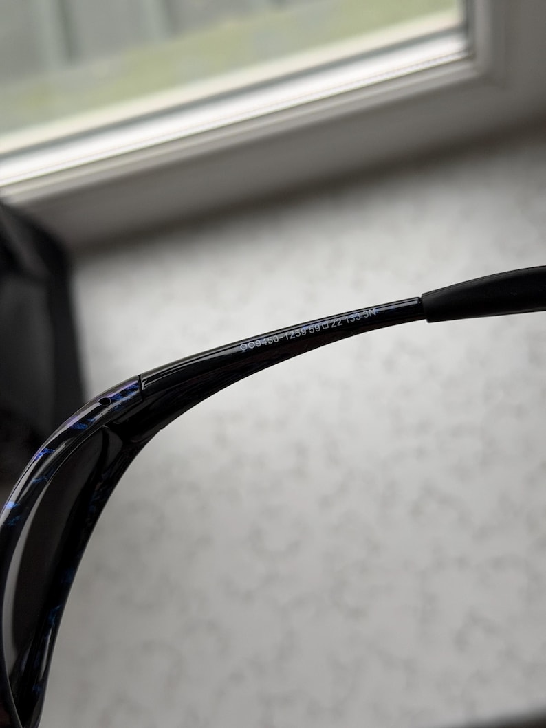 K&ouml;nnte beinhalten: Schwarze Sonnenbrille mit geschwungenem Rahmen und blau-schwarzem Muster auf der Innenseite des Rahmens. Der Text "G09460-1259 59 22 135 N" ist auf dem B&uuml;gel aufgedruckt.