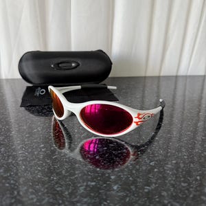 May include: White sunglasses with red lenses and a flame design on the temples. The sunglasses are accompanied by a black case and a cleaning cloth. The lenses reflect the surrounding environment. The sunglasses are on a dark, textured surface.