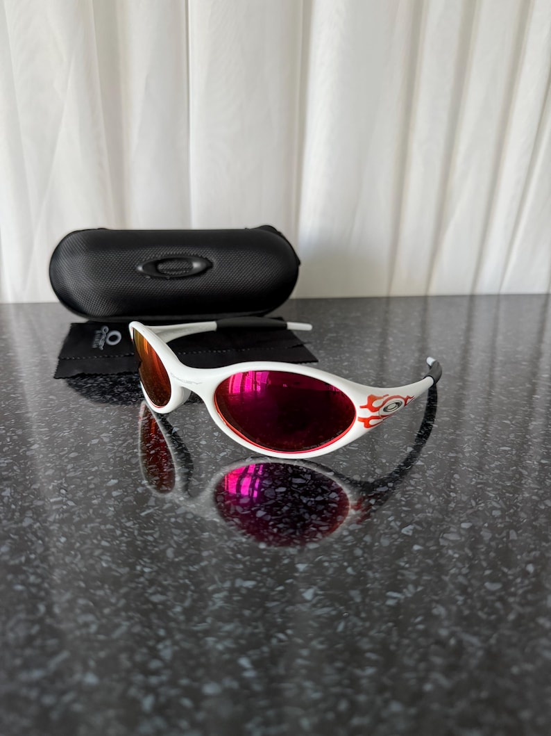 May include: White sunglasses with red lenses and a flame design on the temples. The sunglasses are accompanied by a black case and a cleaning cloth. The lenses reflect the surrounding environment. The sunglasses are on a dark, textured surface.